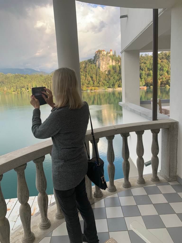 The author of Travel The Four Corners, a travelogue, is photographed taking a photo of Lake Bled from Grand Hotel Toplice in Slovenia.
